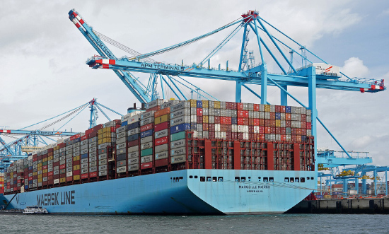 A blue Maersk container shipwith hundreds and hundreds of cargo containers on its deck, with more being loaded by cranes